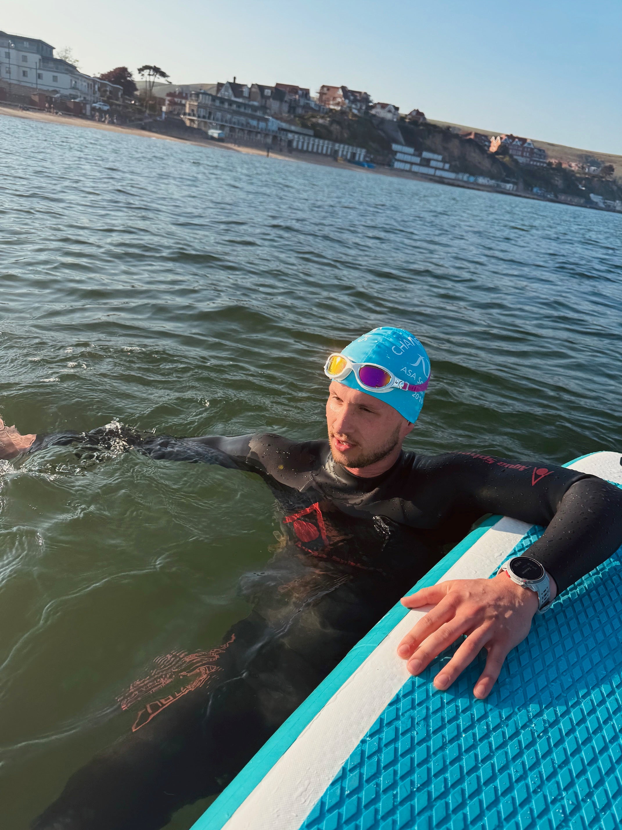 Man in a swimming wetsuit and swim cap next to a paddleboard in the water with a coastal background.
